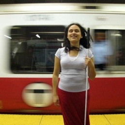 Lindsay Yazzolino, a woman with shoulder length brown hair holding a white cane and standing on a platform in the subway station as the redline subway train zips by behind her.