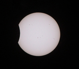 The Sun appears as a glowing, white disk against a black background. On the left side of the Sun, a small, U-shaped area appears missing from the Sun’s perfect circle shape. This missing area is the Moon as it begins to slide in front of the Sun. The Moon appears completely black and smooth.