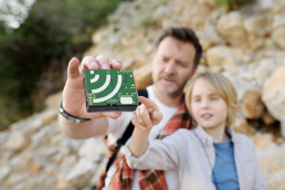 Man holding an AudioMoth recording device out in front of himeself as he shows it to a younger person while outside in nature..