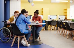 A diverse group of students, including one in a wheelchair, studying together at table in college computer lab