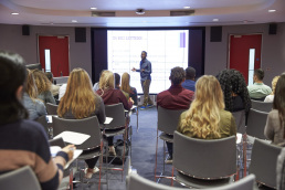 Student lecture in modern university classroom, with lecturer in front of a wall of video screens.