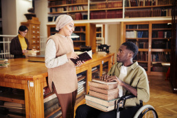 A young African-American man in wheelchair holding stack of books and chatting with a person holding a book at a library
