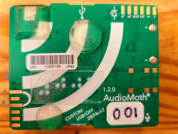 AudioMoth device which is a small square about hald the size of a smart phone. It is eqiuiped with nump dots and in the bottom right corner the numbers 001 are printed on the device.