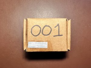 Small square cardboard box with the number 001 written in large print and a braille label attched in the bottom left corner.