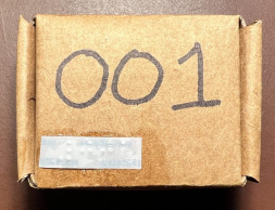 Small square cardboard box with the number 001 written in large print and a braille label attched in the bottom left corner.