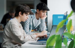 two white male presenting students working on computers side by side in a classroom setting