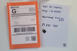 rectangular return mailer on a desk next to a piece of paper with latitude, longitude, start time, and ES ID number and a MicroSD card.