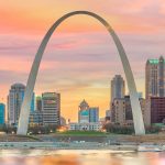 Sunset view of the St. Louis Gateway Arch framing downtown buildings, with the Old Courthouse centered beneath a colorful pink and orange sky and the river in the foreground.