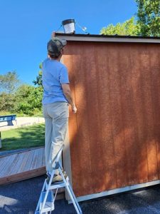 A person standing on a small step ladder beside a wooden shed, reaching up to adjust equipment mounted near the roof on a sunny day.
