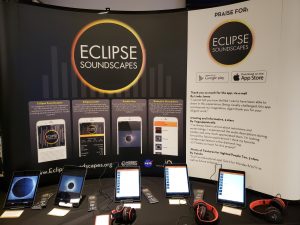 A conference booth display for “Eclipse Soundscapes” featuring a large backdrop with the project’s logo and images of the mobile app. In front of the display, several tablets are arranged on a table, each showing different screens from the Eclipse Soundscapes app. A pair of headphones accompanies each tablet for listening to audio content. A second panel on the right highlights user praise for the app.